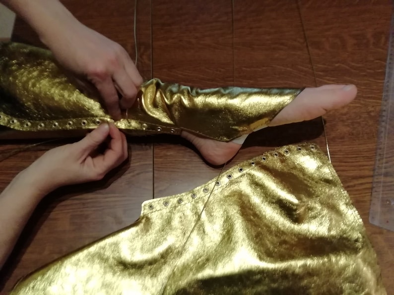Peut inclure: Une paire de jambi&egrave;res en tissu dor&eacute; m&eacute;tallis&eacute; dot&eacute;es d'une s&eacute;rie d'&oelig;illets m&eacute;talliques argent&eacute;s sur les bords lat&eacute;raux. Le tissu pr&eacute;sente une surface textur&eacute;e et r&eacute;fl&eacute;chissante, con&ccedil;ue pour envelopper le bas de la jambe avec un la&ccedil;age.