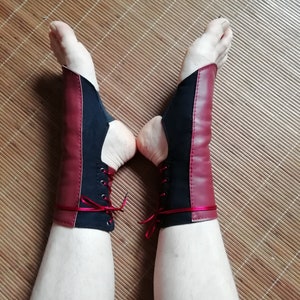 May include: A pair of ankle wraps featuring a combination of black fabric and deep red panels. The wraps include a lace-up design with metal eyelets and thin red ribbon laces that tie around the lower leg, leaving the heel and toes exposed.