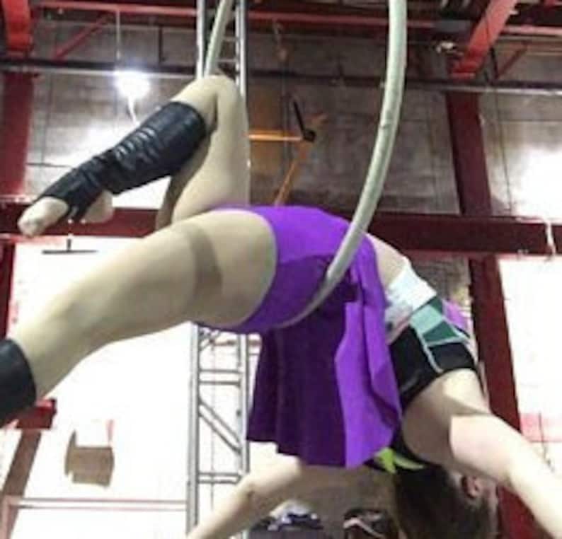 Peut inclure: Un artiste portant une jupe violette et un haut de sport noir avec des accents verts, ainsi que des jambi&egrave;res noires, ex&eacute;cute une pose sur un cerceau a&eacute;rien blanc. La personne est suspendue dans les airs, v&ecirc;tue d'une tenue de sport.