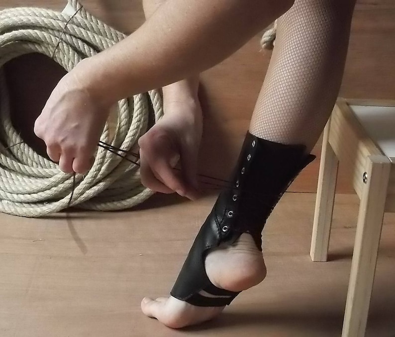 May include: A black leather ankle wrap featuring a lace-up design with silver-toned metal eyelets. The accessory covers the front of the ankle and the top of the foot, secured with thin black laces that wrap around the lower leg for a snug fit.