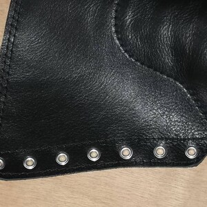 May include: A black leather panel featuring a straight edge lined with seven evenly spaced silver-toned metal eyelets. The material has a visible grain texture and is secured with reinforced stitching along the perimeter of the eyelet strip.