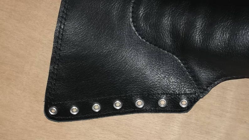 May include: A black leather panel featuring a straight edge lined with seven evenly spaced silver-toned metal eyelets. The material has a visible grain texture and is secured with reinforced stitching along the perimeter of the eyelet strip.