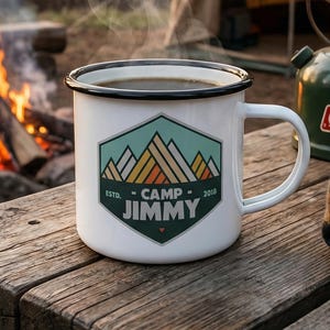 May include: A white enamel mug with a black rim and the words "Camp Jimmy" printed on it, filled with coffee, sits on a wooden table. A campfire burns in the background, with a green Coleman lantern and a plaid blanket nearby. The scene evokes a camping theme.