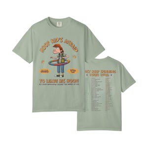 May include: A sage green t-shirt featuring a cartoon character with a hula hoop and the text "HOOP KID'S AFRAID TO LEAVE HIS HOOP!" on the front. The back displays "JUST KEEP SPINNING TOUR 2026" with a list of tour dates and locations printed in black.