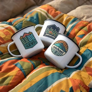 May include: Three white enamel mugs with black rims and handles, each featuring a unique mountain and nature-themed design. One mug has "LEVI" printed on it, another "Jameson", and the third "michael". The mugs are arranged on a colorful patterned blanket.
