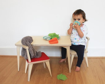 Puede incluir: Un niño se sienta en un juego de mesa y silla de madera con un conejito de peluche en la otra silla. La mesa tiene un plato con una verdura verde de hoja y dos zanahorias naranjas encima. El niño sostiene una taza azul y blanca.