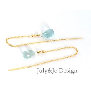 Puede incluir: Pendientes colgantes dorados con gemas en forma de lágrima de color azul claro. Los pendientes tienen una cadena y una barra recta. El texto "July&Jo Design" es visible.