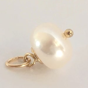 May include: A close-up of a pearl pendant with a gold bail and a small gold bead on top. The pearl is a creamy white color and has a smooth, lustrous surface. The pendant is likely for a necklace or bracelet.