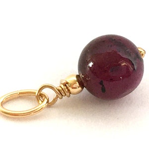 Può includere: Un ciondolo d'oro con una pietra preziosa rotonda di colore rosso scuro. Il ciondolo ha un piccolo anello d'oro per attaccarlo a una collana o un braccialetto.