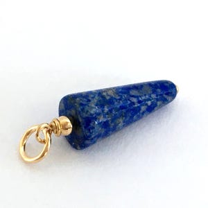 May include: A gold pendant featuring a blue lapis lazuli stone shaped like a cone. The stone has a speckled pattern and is attached to a gold loop.