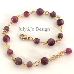 May include: A gold-toned chain bracelet featuring alternating round and irregular-shaped, deep red gemstones. The bracelet has a lobster clasp closure. The text "July&Jo Design" is visible on the image.