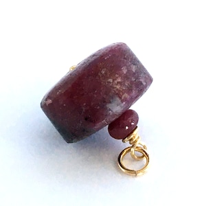 Op de afbeelding: Een bordeauxrode edelsteen hanger met een rechthoekige hoofdsteen en een kleinere, ronde steen eronder. De stenen zijn verbonden met goudkleurig draad en een kleine gouden ring. De hanger staat tegen een witte achtergrond.