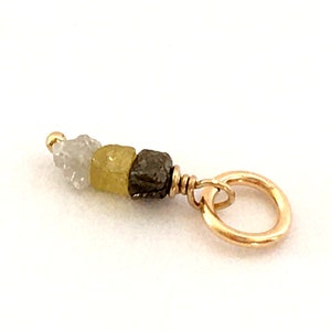 May include: A gold charm featuring three uncut gemstones in grey, yellow, and dark grey. The charm has a small gold bead at the top and a gold ring at the bottom for attachment.