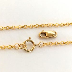 以下が含まれることがあります： ゴールドカラーのネックレスチェーンのクローズアップ。ロブスタークラスプとスプリングリングクラスプが付いています。チェーンは小さな楕円形のリンクでできています。留め金もゴールドカラーです。