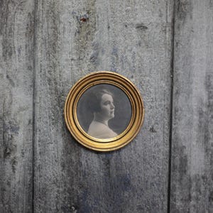 Peut inclure: Un portrait circulaire vintage dans un cadre doré. L'image en noir et blanc représente une femme de profil. Le cadre est posé sur un fond en bois gris vieilli, créant une esthétique rustique.