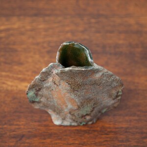 K&ouml;nnte beinhalten: Handgefertigte, kleine Keramikvase mit rauer, strukturierter Oberfl&auml;che. Die Vase ist haupts&auml;chlich braun mit gr&uuml;nen Akzenten und einem dunkelgr&uuml;nen, steinartigen Element in der Mitte. Sie steht auf einer Holzoberfl&auml;che.