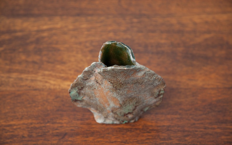 K&ouml;nnte beinhalten: Handgefertigte, kleine Keramikvase mit rauer, strukturierter Oberfl&auml;che. Die Vase ist haupts&auml;chlich braun mit gr&uuml;nen Akzenten und einem dunkelgr&uuml;nen, steinartigen Element in der Mitte. Sie steht auf einer Holzoberfl&auml;che.