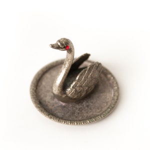 May include: A silver swan figurine with red eyes sits on a silver tray. The swan has detailed feathers and a delicate beak.