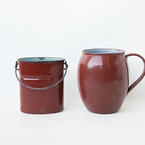 Op de afbeelding: Twee rode emaille containers, een met een handvat en de andere met een metalen handvat. Beide containers hebben een lichtblauwe rand.