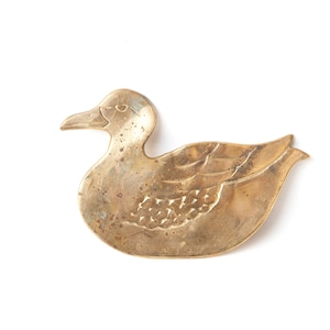 May include: A gold-colored metal duck figurine. The duck is depicted in a swimming pose with its wings spread out. The figurine has a detailed design with a textured surface.