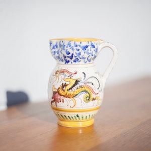 May include: A white ceramic pitcher with a yellow, blue, and red dragon design. The pitcher has a handle and a wide mouth.