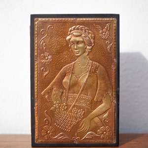 May include: A vintage wooden box with a copper-colored embossed image of a woman with a floral design. The woman is wearing a long dress and a necklace. The box has a black frame.