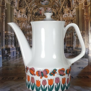 Peut inclure: Théière en céramique blanche avec un motif floral composé de tulipes orange et de papillons roses. La théière a une base en bois.