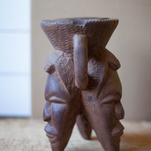 Op de afbeelding: Een houten sculptuur van twee gezichten met een gegraveerde handgreep. De sculptuur is bruin en heeft een getextureerd oppervlak.