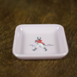 May include: A pink square ashtray with a white outline and a walking man graphic in the center. The graphic includes the text "Johnnie Walker" and "Keep Walking".