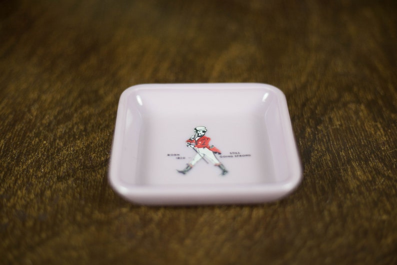 May include: A pink square ashtray with a white outline and a walking man graphic in the center. The graphic includes the text "Johnnie Walker" and "Keep Walking".