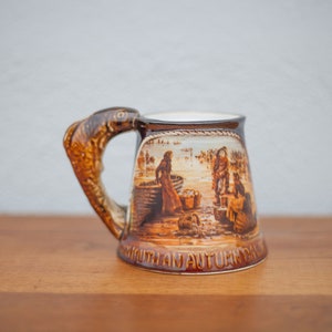Peut inclure: Une tasse en céramique brune avec une anse en forme de poisson. La tasse représente une scène de personnes pêchant dans un bateau avec le texte "Southam Autumn Cal"