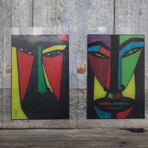 May include: Two abstract art prints featuring stylized faces with bold colors. One face has red and green accents, the other has purple, blue, and green. Both are set against a black background with yellow borders.