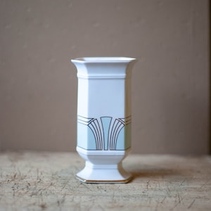 Op de afbeelding: Een witte keramische vaas met een geometrisch ontwerp in groen en goud. De vaas heeft een vierkante basis en een cilindrisch lichaam.