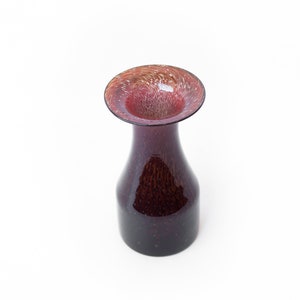 May include: A small, dark red glass vase with a wide, flared opening. The vase has a speckled, textured surface and a swirling pattern in the opening.