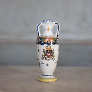 May include: A white ceramic vase with two handles, featuring a colorful design. The vase has a yellow base and is adorned with a crest and floral patterns. The vase is approximately 6 inches tall.