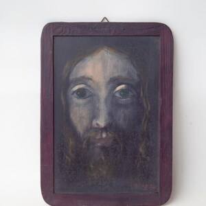 May include: A framed painting of a man's face with a beard. The painting is done in a dark, muted color palette and the man's eyes are looking directly at the viewer. The frame is a dark wood with a purple tint.
