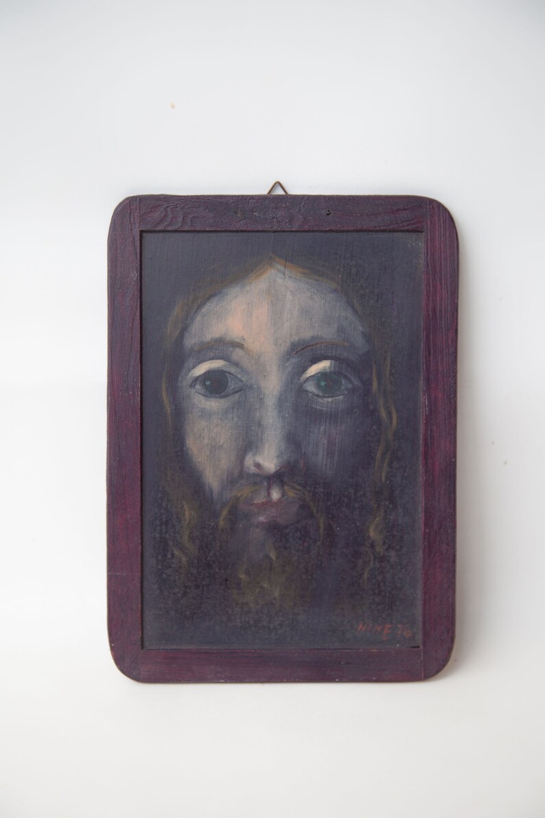 May include: A framed painting of a man's face with a beard. The painting is done in a dark, muted color palette and the man's eyes are looking directly at the viewer. The frame is a dark wood with a purple tint.