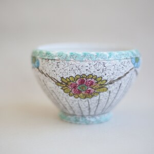 May include: A white ceramic bowl with a blue and green rim and floral designs. The bowl has a speckled glaze and a scalloped edge.