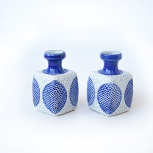 Op de afbeelding: Een paar blauwe en witte keramische kaarsenhouders met een geometrisch ontwerp. De houders zijn vierkant met een ronde opening aan de bovenkant.