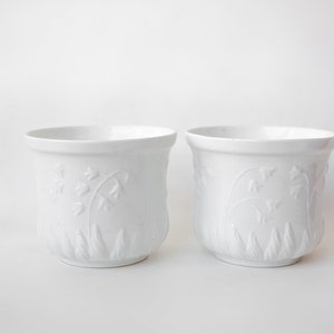 May include: Two white ceramic flower pots with a raised design of lily of the valley flowers and leaves.