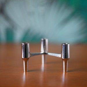Op de afbeelding: Een zilverkleurige kandelaar met drie armen. Elke arm heeft een cilindrische beker voor het vasthouden van een kaars. De kandelaar is gemaakt van metaal en heeft een modern, minimalistisch design. De basis heeft een driepootvorm.