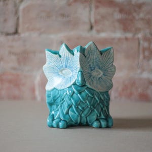 Può includere: Una statuetta di gufo in ceramica color turchese con finitura lucida e dettagli di piume testurizzati sul corpo. Il gufo presenta grandi zone oculari stilizzate con motivi a linee radianti bianche e blu e un piccolo becco appuntito.