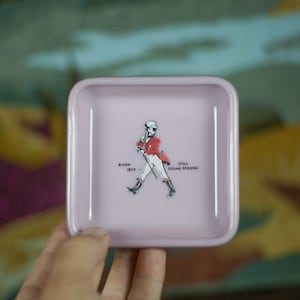 Op de afbeelding: Een roze vierkante sieradenbak met een witte rand. De bak heeft een cartoonillustratie van een man in een rood jasje en witte broek die met een stok loopt. De tekst "Born 1820" en "Still Going Strong" staat onder de man.