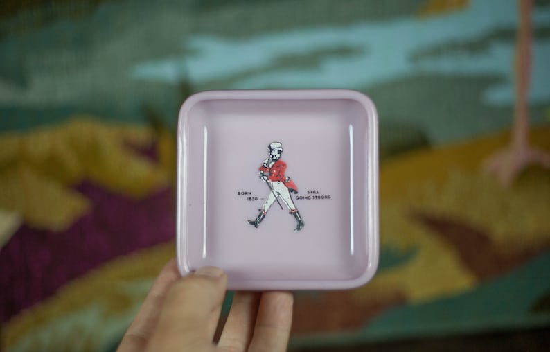 May include: A pink square-shaped trinket tray with a white outline. The tray features a cartoon illustration of a man in a red jacket and white pants walking with a cane. The text "Born 1820" and "Still Going Strong" is printed below the man.