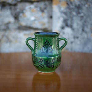 Può includere: Un vaso in ceramica verde con disegni floreali neri e due manici. Il vaso è appoggiato su una superficie di legno.