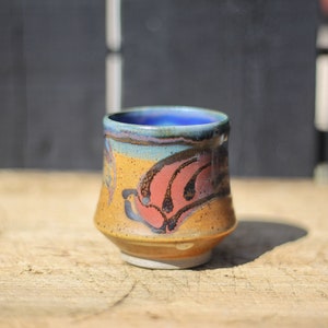 Puede incluir: Una taza de cerámica pequeña pintada a mano con un diseño floral azul, amarillo y rojo. La taza tiene un fondo redondeado y un borde ligeramente acampanado.