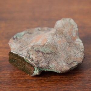 K&ouml;nnte beinhalten: Ein unregelm&auml;&szlig;ig geformtes Objekt mit verwittertem Aussehen. Die Oberfl&auml;che zeigt eine Mischung aus Braun-, Gr&uuml;n- und Kupfert&ouml;nen, m&ouml;glicherweise durch Oxidation. Die Textur ist rau und uneben, mit einer deutlichen Kante, was auf ein dekoratives Objekt hindeutet.