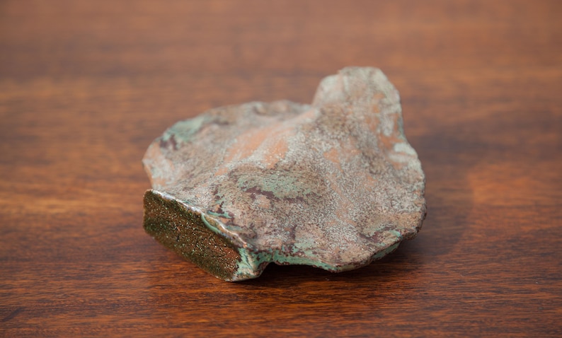 K&ouml;nnte beinhalten: Ein unregelm&auml;&szlig;ig geformtes Objekt mit verwittertem Aussehen. Die Oberfl&auml;che zeigt eine Mischung aus Braun-, Gr&uuml;n- und Kupfert&ouml;nen, m&ouml;glicherweise durch Oxidation. Die Textur ist rau und uneben, mit einer deutlichen Kante, was auf ein dekoratives Objekt hindeutet.