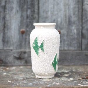 May include: White textured ceramic vase with two green fish designs. The vase has a rounded shape and a narrow opening. The background is a weathered wooden door.