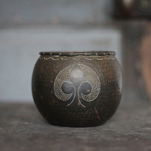 May include: A round, dark brown decorative bowl with a lid. The bowl has a textured surface and a silver-coloured, stylized leaf design on the front. The rim of the bowl is also decorated with a silver pattern. The bowl is on a dark surface.